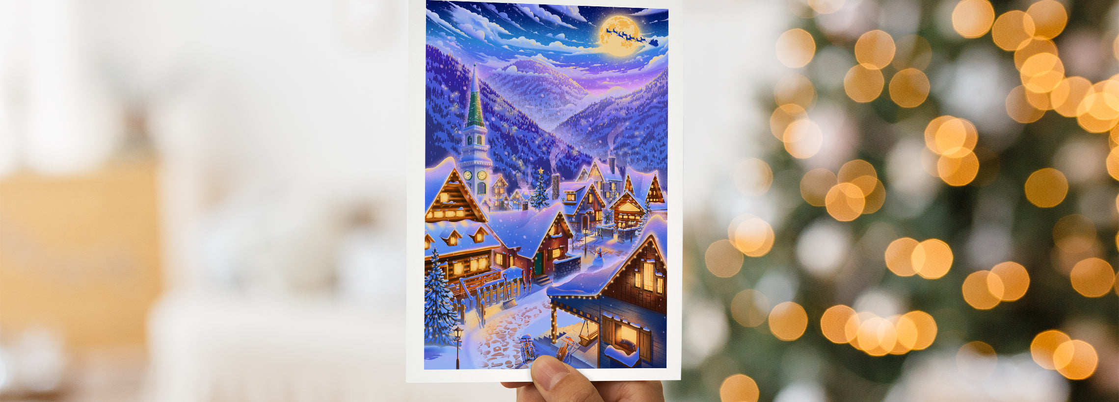 A hand holds a greeting card in front of an out-of-focus lit tree. The card is predominantly blue,  and features a small town in the snow at night, with warm incandescent light spilling from the windows. In the far distance are mountains, with Santa and reindeer crossing a full moon.